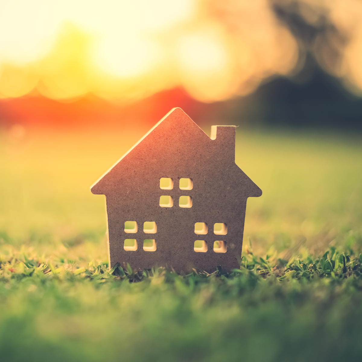 A small brown wooden house-shaped ornament with cutout windows stands on green grass against a warm blurred sunset background, with a light blue wave graphic at the bottom.