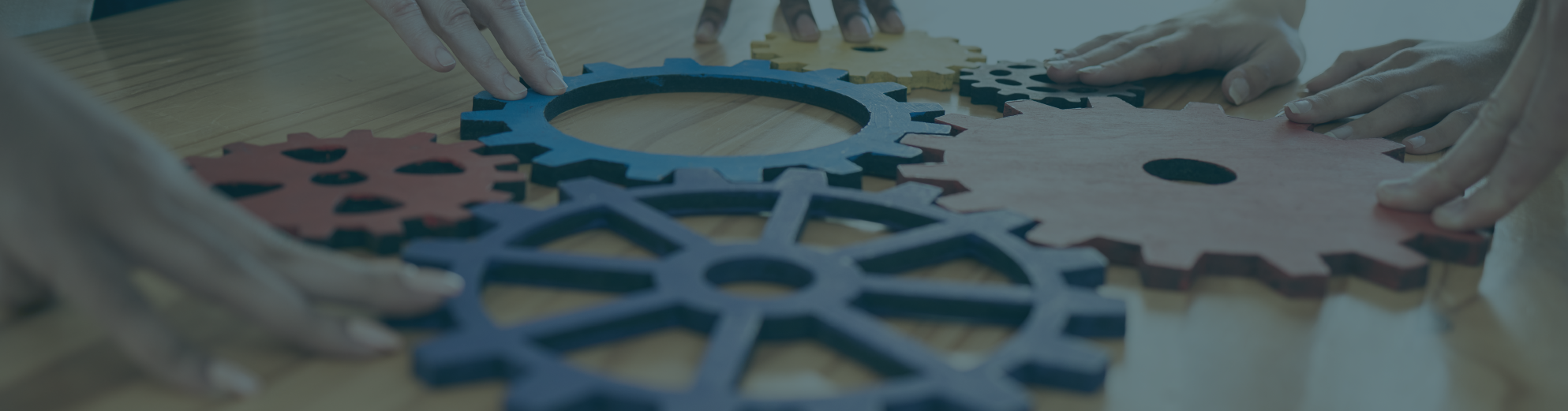 Several hands assembling colorful interlocking gear puzzle pieces on a wooden table, with a blue translucent overlay covering the scene.