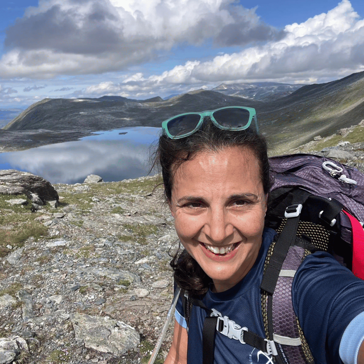 A smiling woman taking a selfie outdoors on a mountain trail, wearing a navy blue shirt, a purple hiking backpack, and green sunglasses, with a scenic lake and rocky landscape in the background.