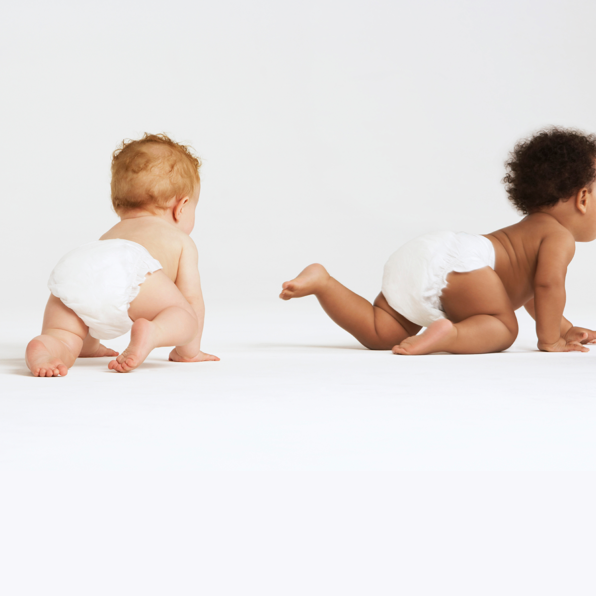 Two babies wearing white diaper cover outfits crawling on a white studio floor against a white background, with a teal and white wavy graphic at the bottom.