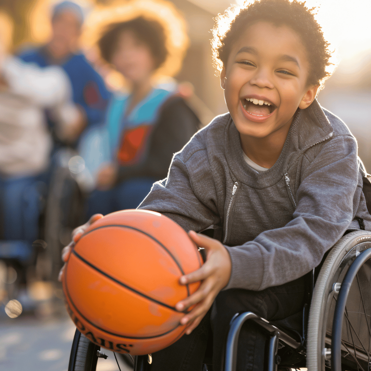 A young boy with curly hair smiling and laughing while holding an orange basketball in a wheelchair. The background shows a softly blurred outdoor scene with other people in wheelchairs, creating a warm, supportive atmosphere.