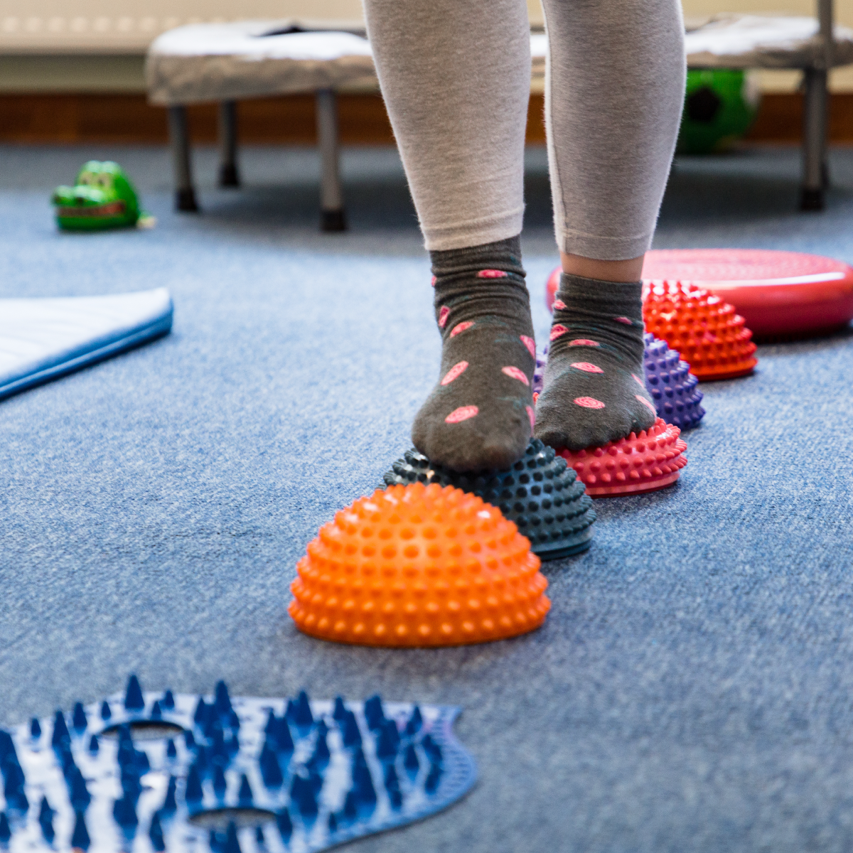 A person wearing gray socks with pink polka dots balances on a row of colorful textured dome exercise balls on a blue carpeted floor.