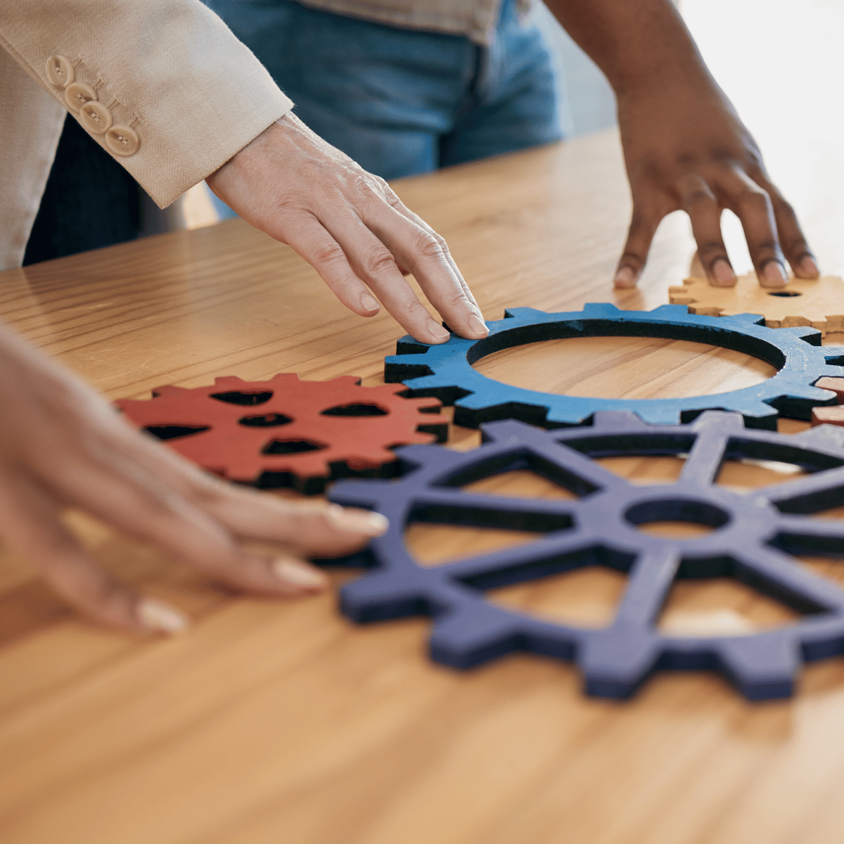 Three interlocking wooden gear puzzle pieces in blue, dark navy, and red on a light wood table, with multiple hands adjusting them. A person wearing a beige blazer and blue jeans is partially visible in the background.